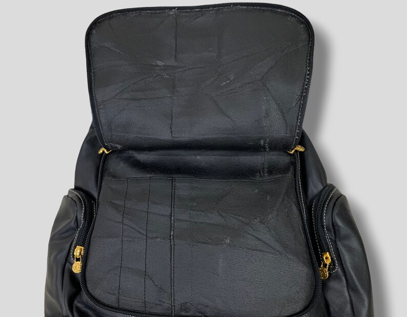 May include: Black leather backpack with an open top, revealing the interior compartments. The backpack features gold-colored zippers and white stitching details. The design includes multiple pockets and a structured shape, suitable for carrying various items.