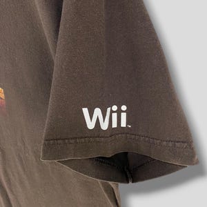 May include: A brown t-shirt featuring the word "Wii" in white on the sleeve. The shirt is made of a soft material and has a classic crew neck design. The sleeve is short and the shirt is likely a casual wear item.