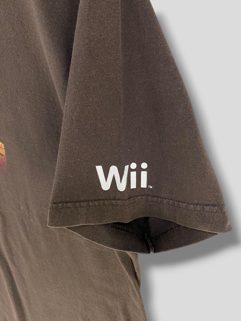 May include: A brown t-shirt featuring the word "Wii" in white on the sleeve. The shirt is made of a soft material and has a classic crew neck design. The sleeve is short and the shirt is likely a casual wear item.