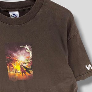 May include: A brown t-shirt with a vibrant graphic print. The print depicts a fantasy scene with figures, a bright light, and a circular design. The sleeve has a white letter "W".
