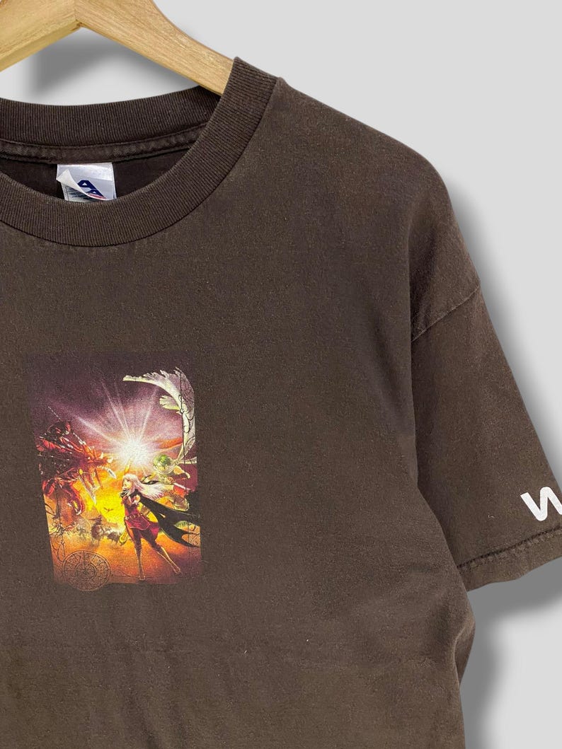 May include: A brown t-shirt with a vibrant graphic print. The print depicts a fantasy scene with figures, a bright light, and a circular design. The sleeve has a white letter "W".