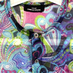 Puede incluir: Una blusa transparente con botones y un vibrante estampado de cachemira en tonos de azul, morado, verde y rosa. La blusa presenta un cuello mandar&iacute;n y la marca "tricot COMME des GAR&Ccedil;ONS" en una etiqueta negra.