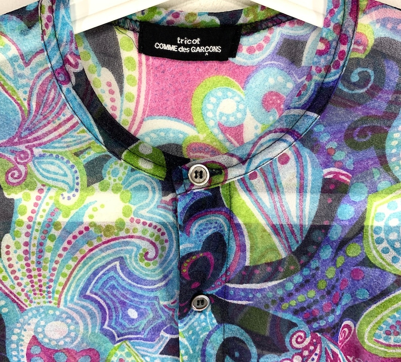 Puede incluir: Una blusa transparente con botones y un vibrante estampado de cachemira en tonos de azul, morado, verde y rosa. La blusa presenta un cuello mandar&iacute;n y la marca "tricot COMME des GAR&Ccedil;ONS" en una etiqueta negra.