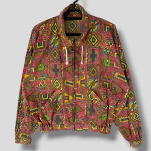 Op de afbeelding: Een vintage jas met ritssluiting en een levendig geometrisch patroon in tinten rood, geel, groen en blauw. De jas heeft een hoge kraag, een capuchon met trekkoord en twee voorzakken. De jas hangt aan een zwarte hanger.