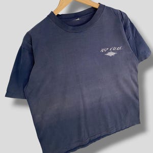 May include: A faded navy blue t-shirt with the text "RIP CURL" in white. The shirt is short-sleeved and has a crew neck. The Rip Curl logo is printed on the upper left chest area of the shirt.