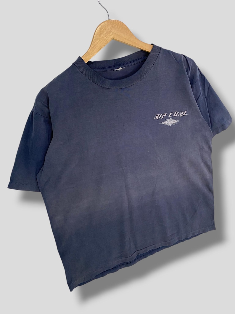 May include: A faded navy blue t-shirt with the text "RIP CURL" in white. The shirt is short-sleeved and has a crew neck. The Rip Curl logo is printed on the upper left chest area of the shirt.