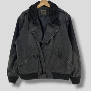 Könnte beinhalten: Schwarze Bomberjacke mit einem Kunstfellkragen. Die Jacke hat einen Reißverschluss, mehrere Taschen und gerippte Bündchen und Saum. Die Jacke hängt an einem Holzbügel.