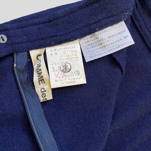Puede incluir: Primer plano de una prenda azul marino con cremallera y etiqueta. La etiqueta dice "COMME des GAR&Ccedil;ONS" e incluye texto japon&eacute;s con detalles de la composici&oacute;n de la tela. La prenda parece ser de lana y nailon.