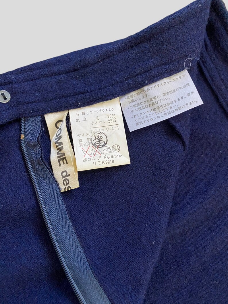 Puede incluir: Primer plano de una prenda azul marino con cremallera y etiqueta. La etiqueta dice "COMME des GAR&Ccedil;ONS" e incluye texto japon&eacute;s con detalles de la composici&oacute;n de la tela. La prenda parece ser de lana y nailon.