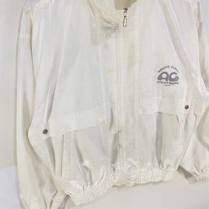 K&ouml;nnte beinhalten: Eine wei&szlig;e, leichte Jacke mit hohem Kragen und durchgehendem Rei&szlig;verschluss. Die Jacke hat zwei Vordertaschen und elastische B&uuml;ndchen. Der Text "sport futur courr&egrave;ges paris" ist auf der Brust aufgedruckt. Die Jacke ist aus gl&auml;nzendem Material.