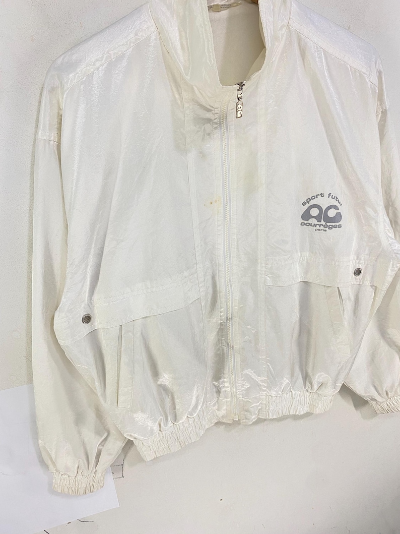 K&ouml;nnte beinhalten: Eine wei&szlig;e, leichte Jacke mit hohem Kragen und durchgehendem Rei&szlig;verschluss. Die Jacke hat zwei Vordertaschen und elastische B&uuml;ndchen. Der Text "sport futur courr&egrave;ges paris" ist auf der Brust aufgedruckt. Die Jacke ist aus gl&auml;nzendem Material.