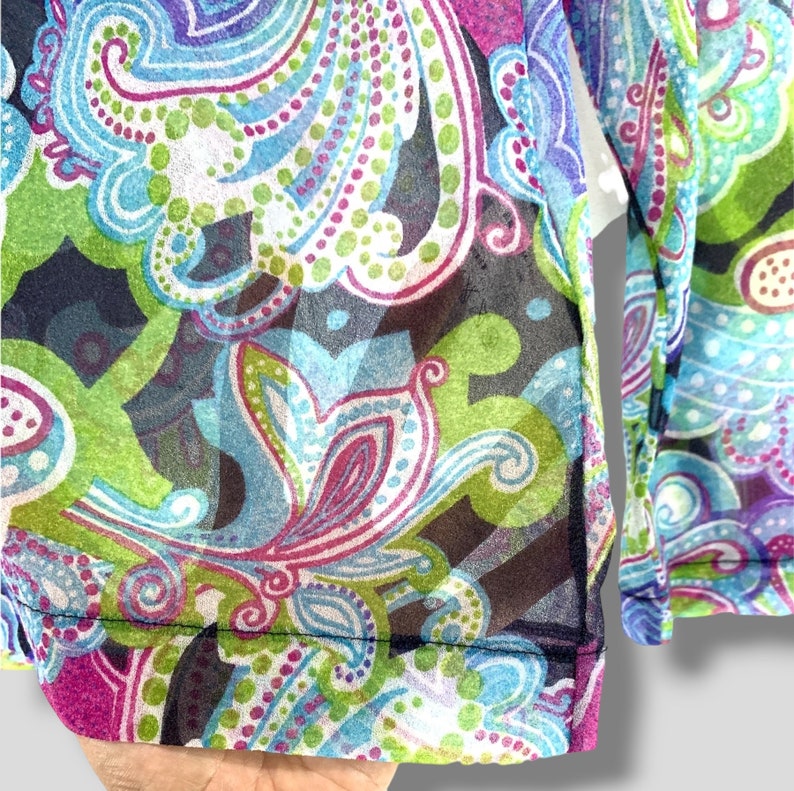 Puede incluir: Una blusa transparente de manga larga con un vibrante estampado de cachemira en tonos azules, verdes, morados y rosas. La tela es ligera y semitransparente, con un ribete azul marino oscuro a lo largo del dobladillo y los pu&ntilde;os.