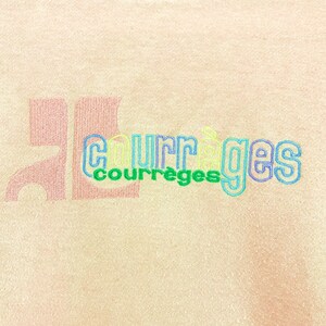 May include: Close-up of a light peach-colored fabric with the embroidered text "COURR&Egrave;GES" in a multi-colored, outlined font. A large, stylized "JL" is printed in a darker peach color to the left of the text.