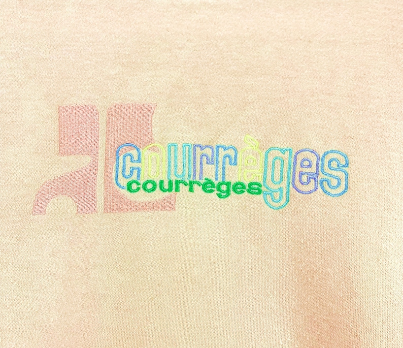 May include: Close-up of a light peach-colored fabric with the embroidered text "COURR&Egrave;GES" in a multi-colored, outlined font. A large, stylized "JL" is printed in a darker peach color to the left of the text.