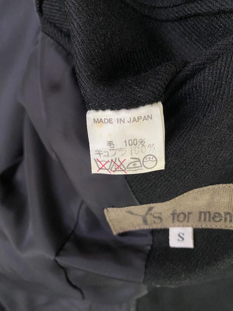 May include: A black blazer with a label that reads "MADE IN JAPAN" and "毛 100% キュプラ 100%". The label also has a care tag with a dry clean symbol and a medium heat iron symbol. The blazer has a label that reads "Y's for men" and "S" for the size.