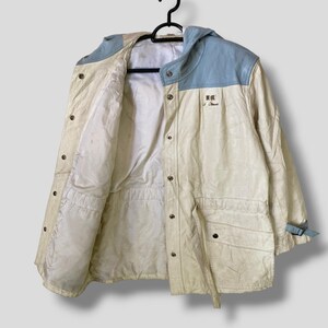 Puede incluir: Una chaqueta blanca con capucha con un panel azul en la capucha y un parche azul en el lado derecho del pecho. La chaqueta tiene una cintura con cord&oacute;n y una cremallera.