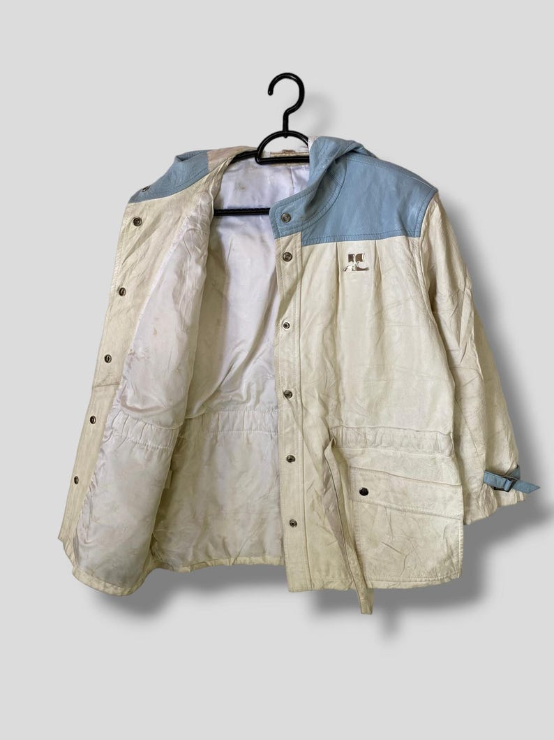 Puede incluir: Una chaqueta blanca con capucha con un panel azul en la capucha y un parche azul en el lado derecho del pecho. La chaqueta tiene una cintura con cord&oacute;n y una cremallera.