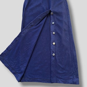 Puede incluir: Una falda azul marino con cierre de botones en la parte delantera. La falda tiene una silueta larga y recta y una abertura dividida en el lado izquierdo, revelando la tela interior. Los botones son oscuros y redondos.