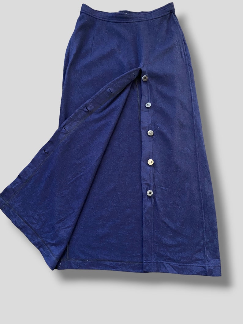 Puede incluir: Una falda azul marino con cierre de botones en la parte delantera. La falda tiene una silueta larga y recta y una abertura dividida en el lado izquierdo, revelando la tela interior. Los botones son oscuros y redondos.