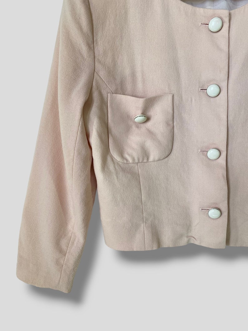 May include: A pale pink blazer with a single patch pocket on the left side. The blazer has a button-up front closure with five white buttons.