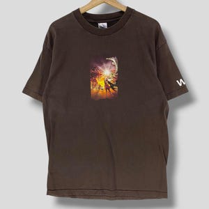 May include: A brown t-shirt with a central graphic print depicting a vibrant, action-filled scene with figures and a bright light. The sleeve has a white letter "w". The shirt is suspended from a wooden hanger.