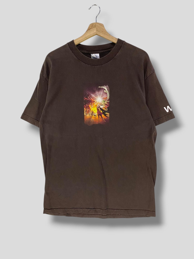 May include: A brown t-shirt with a central graphic print depicting a vibrant, action-filled scene with figures and a bright light. The sleeve has a white letter "w". The shirt is suspended from a wooden hanger.
