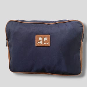 May include: A navy blue rectangular bag with brown trim and a brown leather patch. The patch features a white logo. The bag has a zipper and brown leather accents on the sides.