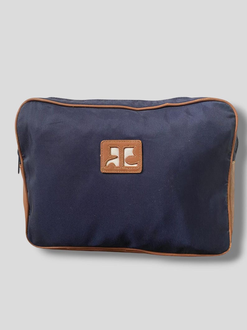 May include: A navy blue rectangular bag with brown trim and a brown leather patch. The patch features a white logo. The bag has a zipper and brown leather accents on the sides.