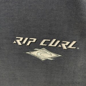 May include: Dark gray fabric with the white text "RIP CURL" and a stylized diamond-shaped logo. The logo features a wave design within a diamond outline. The text and logo appear to be screen printed.