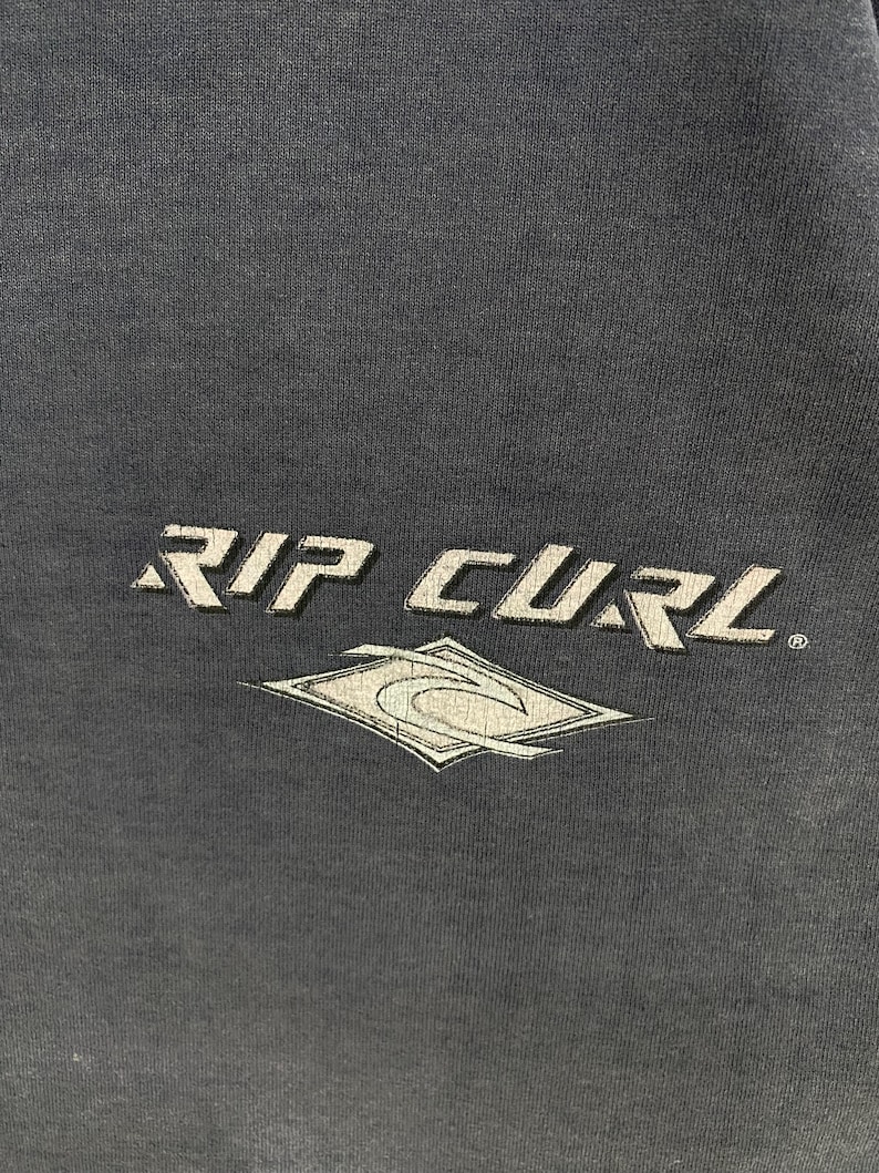 May include: Dark gray fabric with the white text "RIP CURL" and a stylized diamond-shaped logo. The logo features a wave design within a diamond outline. The text and logo appear to be screen printed.