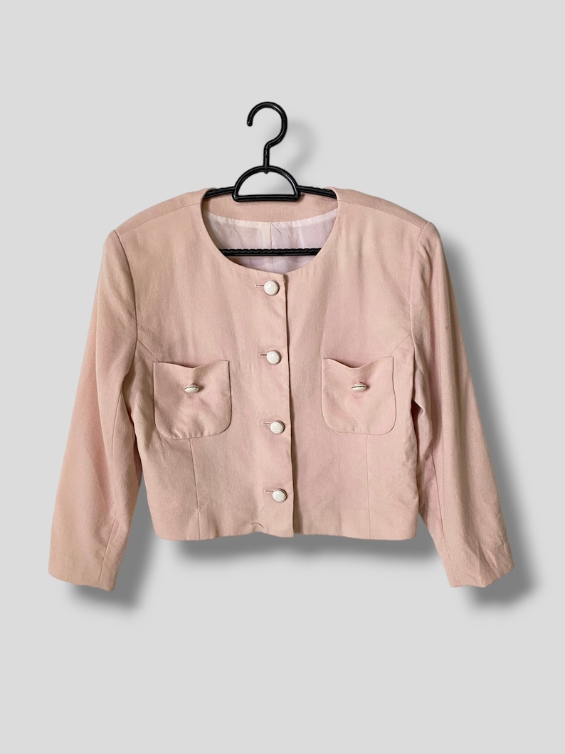 May include: A pink blazer with long sleeves and white buttons. The blazer has two pockets on the front and a round neckline.