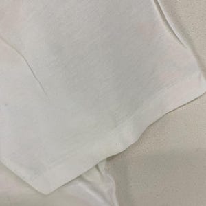 May include: Close-up of a white t-shirt sleeve, showing the fabric's texture and the seam detail. The sleeve is folded, revealing the inside of the garment. The fabric appears to be a lightweight cotton or similar material.