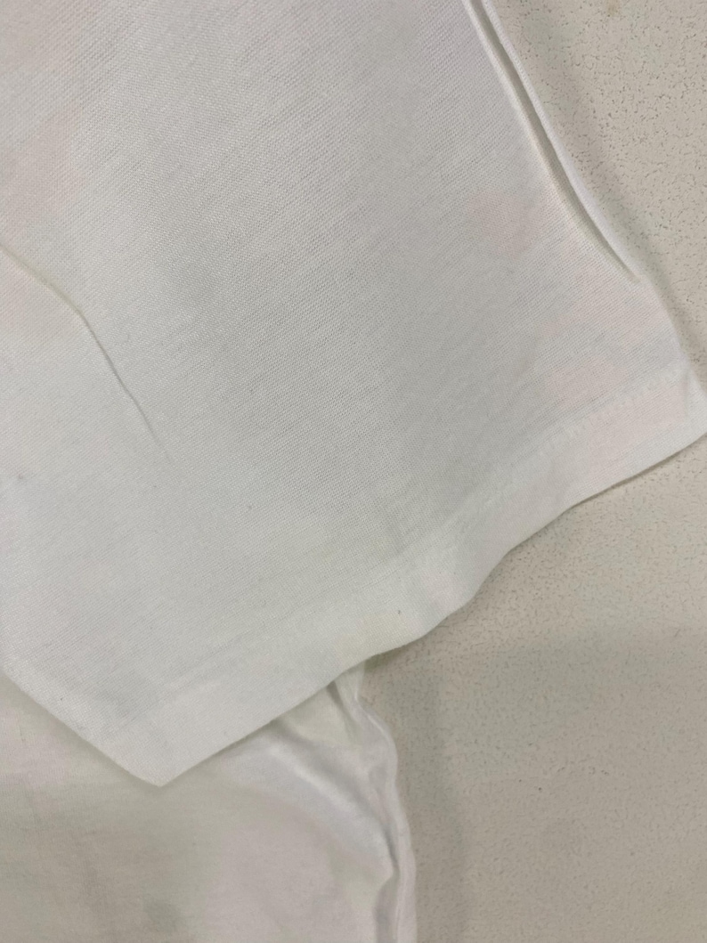 May include: Close-up of a white t-shirt sleeve, showing the fabric's texture and the seam detail. The sleeve is folded, revealing the inside of the garment. The fabric appears to be a lightweight cotton or similar material.