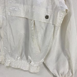 K&ouml;nnte beinhalten: Eine wei&szlig;e Jacke von Courr&egrave;ges Paris. Die Jacke hat einen Rei&szlig;verschluss, eine Tasche und elastische B&uuml;ndchen und Saum. Das Courr&egrave;ges-Logo ist oben links sichtbar. Die Jacke ist aus gl&auml;nzendem, leichtem Material.
