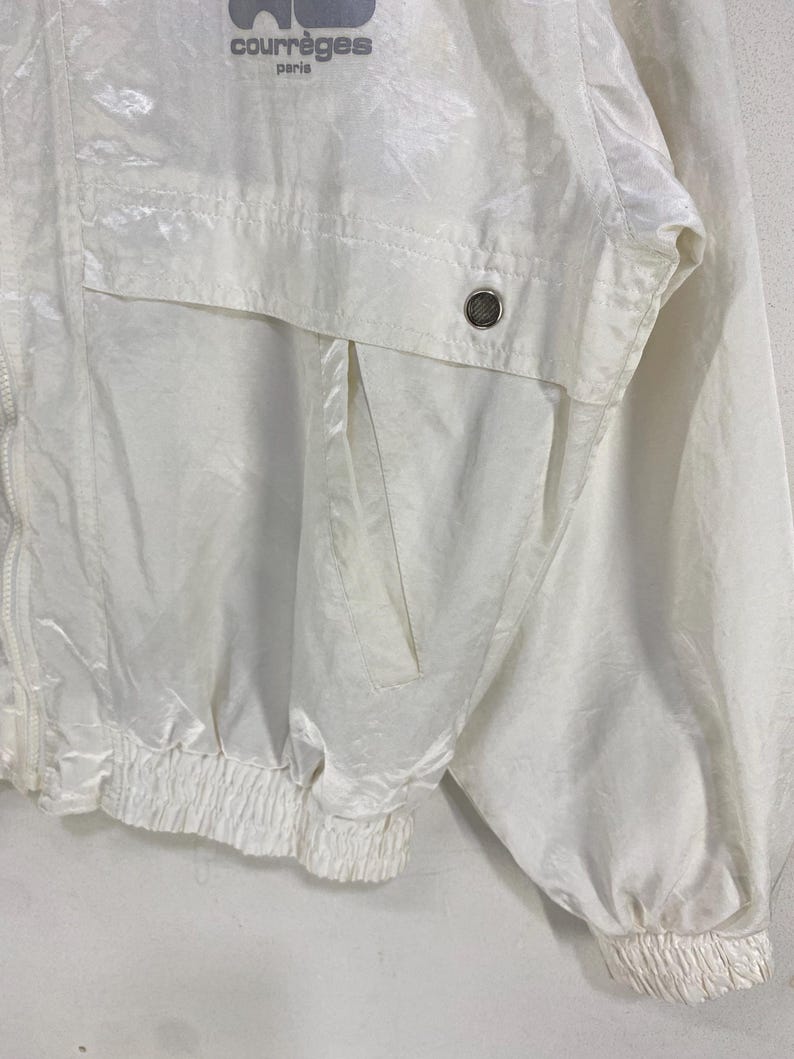 K&ouml;nnte beinhalten: Eine wei&szlig;e Jacke von Courr&egrave;ges Paris. Die Jacke hat einen Rei&szlig;verschluss, eine Tasche und elastische B&uuml;ndchen und Saum. Das Courr&egrave;ges-Logo ist oben links sichtbar. Die Jacke ist aus gl&auml;nzendem, leichtem Material.