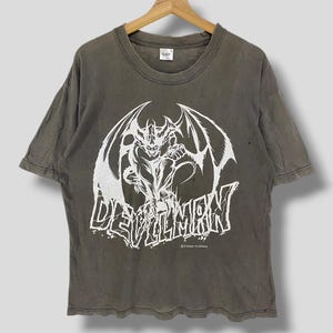 May include: A faded olive green t-shirt featuring a white graphic of a winged demon and the word "DEVILMAN" in a stylized font. The shirt is hanging on a wooden hanger. The graphic includes the text "DYNAMIC PLANNING".