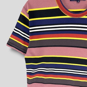 May include: A short-sleeved, striped t-shirt with a crew neck. The shirt features horizontal stripes in various colors, including pink, black, yellow, blue, white, red, and gray. The tag reads "Comme des Garcons" and the size is "M".
