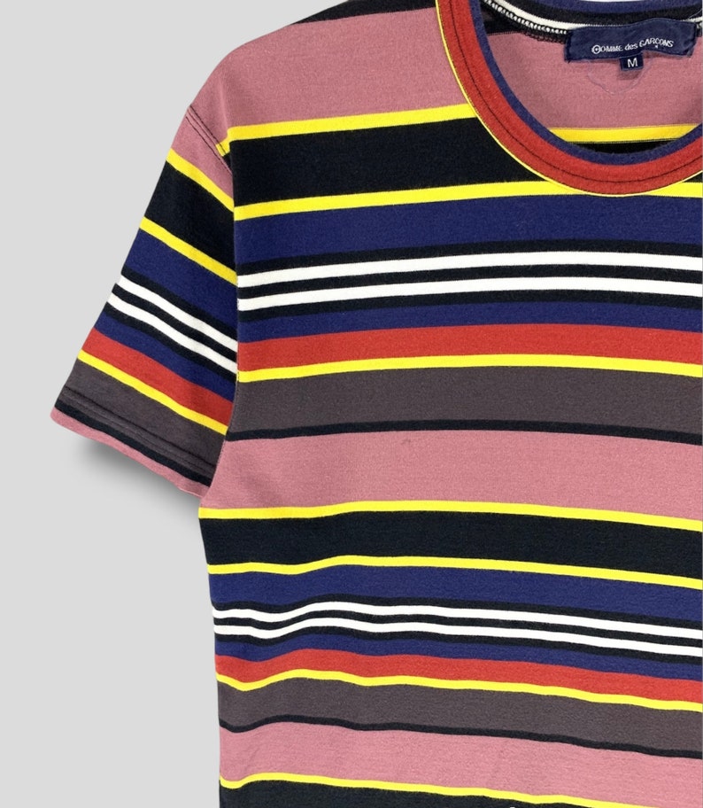 May include: A short-sleeved, striped t-shirt with a crew neck. The shirt features horizontal stripes in various colors, including pink, black, yellow, blue, white, red, and gray. The tag reads "Comme des Garcons" and the size is "M".