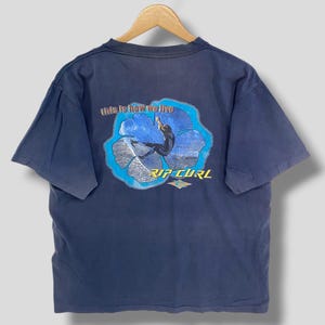 May include: Navy blue t-shirt with a graphic on the back featuring a surfer in a blue and white floral design. The text "this is how we live" is above the surfer, and "RIP CURL" is below.
