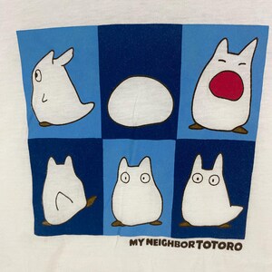 May include: White t-shirt featuring a blue and navy grid design with six cartoon Totoro characters. The text "MY NEIGHBOR TOTORO" is printed in brown at the bottom. The characters are white with brown accents.