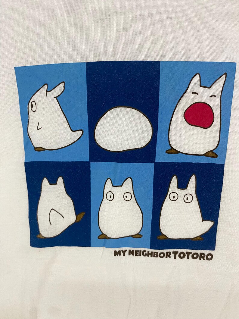 May include: White t-shirt featuring a blue and navy grid design with six cartoon Totoro characters. The text "MY NEIGHBOR TOTORO" is printed in brown at the bottom. The characters are white with brown accents.