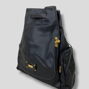 May include: Black leather backpack with gold-toned hardware. Features a drawstring closure, a side zipper pocket, and a small rectangular plaque with gold lettering. White stitching accents the edges.