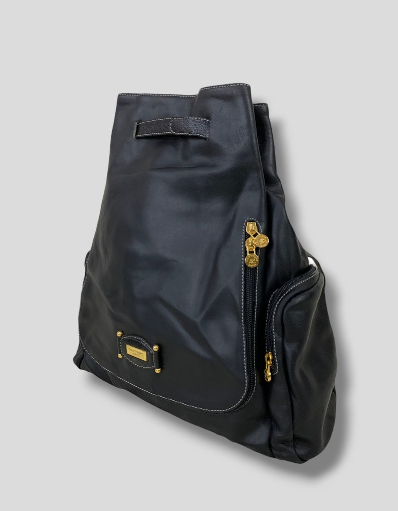 May include: Black leather backpack with gold-toned hardware. Features a drawstring closure, a side zipper pocket, and a small rectangular plaque with gold lettering. White stitching accents the edges.