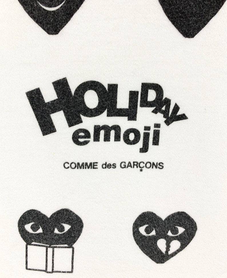 Puede incluir: Tela blanca con corazones gr&aacute;ficos negros y texto. El texto dice "HOLIDAY emoji" encima de "COMME des GAR&Ccedil;ONS". Los corazones tienen varios dise&ntilde;os de ojos y bocas, incluyendo un coraz&oacute;n roto y un coraz&oacute;n leyendo un libro.