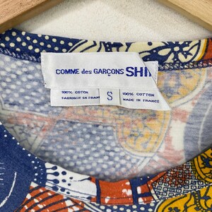 K&ouml;nnte beinhalten: Nahaufnahme eines bunten Shirts mit einem einzigartigen Muster. Das Shirt hat ein Etikett mit der Aufschrift "COMME des GAR&Ccedil;ONS SHIRT", "100% COTON" und "MADE IN FRANCE". Das Shirt hat ein kleines Gr&ouml;&szlig;enetikett, "S". Das Design des Shirts umfasst blaue, orange und gelbe Muster.