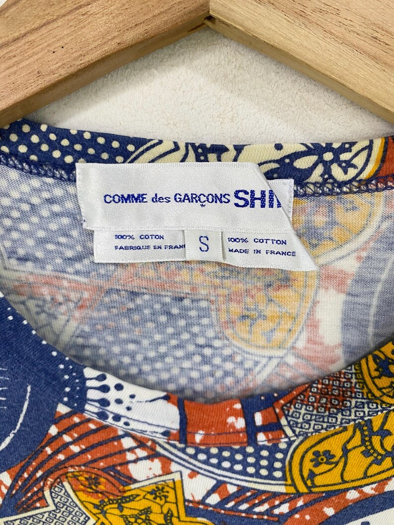 K&ouml;nnte beinhalten: Nahaufnahme eines bunten Shirts mit einem einzigartigen Muster. Das Shirt hat ein Etikett mit der Aufschrift "COMME des GAR&Ccedil;ONS SHIRT", "100% COTON" und "MADE IN FRANCE". Das Shirt hat ein kleines Gr&ouml;&szlig;enetikett, "S". Das Design des Shirts umfasst blaue, orange und gelbe Muster.