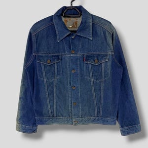 May include: A blue denim jacket with a button closure and two front pockets. The jacket has a faded look and is hanging on a black hanger.