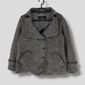 May include: A gray and black herringbone pattern jacket with a wide collar and three buttons. The jacket has two front pockets and is hanging on a black hanger. The jacket's cuffs are folded up.