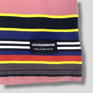May include: Close-up of a striped fabric with a pink top section and horizontal bands of yellow, black, blue, red, and gray. A black label with white text reads "UGO RONDINONE" and "courtesy of Sadie Coles HQ".