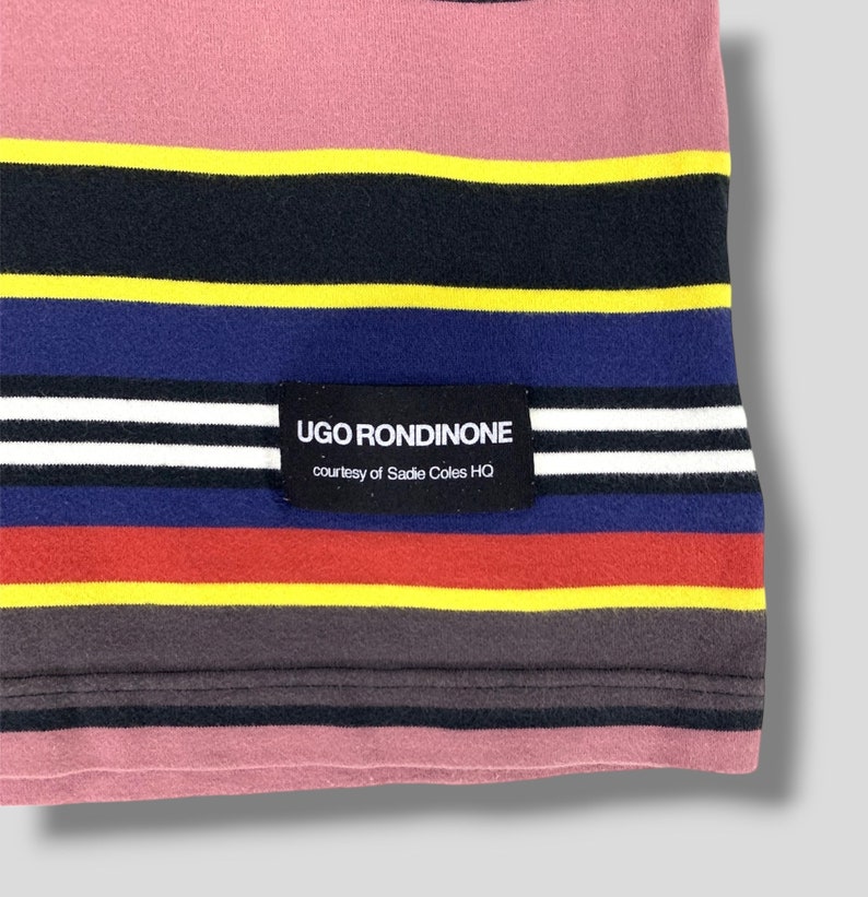 May include: Close-up of a striped fabric with a pink top section and horizontal bands of yellow, black, blue, red, and gray. A black label with white text reads "UGO RONDINONE" and "courtesy of Sadie Coles HQ".